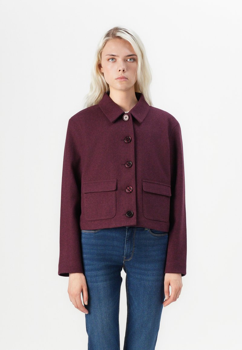 Veste courte bourgogne en tissu texturé, dotée d'un col, d'une fermeture à boutons et de deux poches avant. Portée sur un jean bleu.