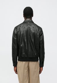 Μαύρο δερμάτινο μπουφάν τύπου bomber με ανάγλυφο κυματιστό σχέδιο, ριμπ τελείωμα και μανσέτες, και κλασικό γιακά. Φορεμένο ανοιχτό στο πίσω μέρος.