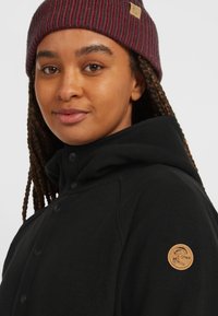 Black fleece jacket with a hood, featuring snap buttons and a circular tan logo patch on the left shoulder. Model wears a burgundy striped beanie.