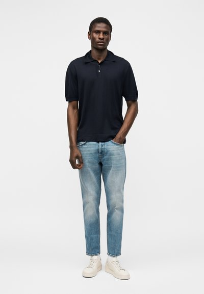 Homme debout avec une main dans la poche, portant un polo noir à manches courtes, un jean bleu clair et des baskets blanches sur un fond uni.