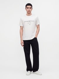 Млад мъж стои с една ръка в джоба, носи бяла тениска Emporio Armani, черен панталон и бели маратонки на едноцветен фон.