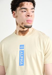 Beige puuvillapaita, jossa on pystysuora sininen grafiikka, jossa lukee "ALPHA" ja tähti-logo. Paidan pyöreä kaula-aukkko, lyhyet hihat ja sileä pinta.
