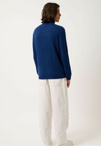 Marineblauer Strickpullover mit geripptem Muster und hohem Kragen, kombiniert mit locker sitzenden cremefarbenen Hosen und weißen Sneakers.