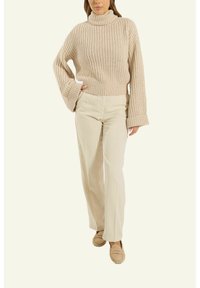 Beige gerippter Strick-Turtleneck-Pullover mit langen, ausgestellten Ärmeln, kombiniert mit hellbeigen geraden Hosen und beige Slippern.