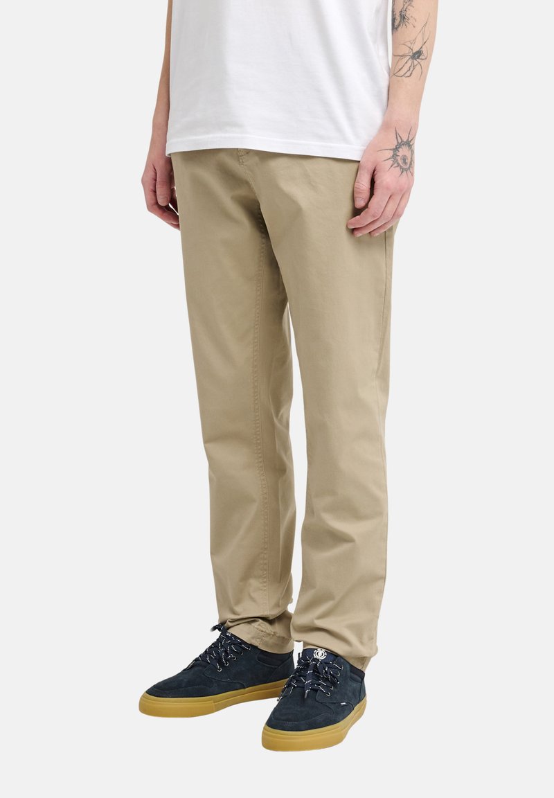 Hombre vistiendo pantalones chinos color beige, zapatillas azul marino con suelas de goma y una camiseta blanca, mostrando tatuajes en el antebrazo y la mano izquierda.