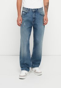 Jeans de mezclilla azul con un corte relajado, que presentan detalles de desgaste y manchas de pintura. Terminados con un cierre de botón y cinco bolsillos.