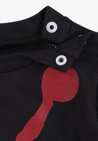 Černá tkanina se dvěma bílými patentkami na límci a červeným tištěným tvarem částečně viditelným na oděvu.