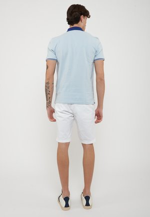 Uomo che indossa una polo azzurro chiaro, pantaloncini bianchi e scarpe da ginnastica bianche, in piedi di spalle, con un tatuaggio floreale sul braccio sinistro.