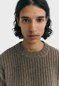 Pull en tricot marron avec un motif texturé et un col rond côtelé. Le modèle a des cheveux bouclés et un fond neutre.