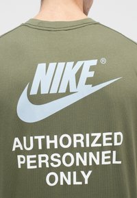 Ελιόχρωμο βαμβακερό T-shirt με μεγάλο μπλε λογότυπο Nike και λευκό κείμενο που γράφει «ΜΟΝΟ ΓΙΑ ΕΓΚΕΚΡΙΜΕΝΟ ΠΡΟΣΩΠΙΚΟ» στο πίσω μέρος.