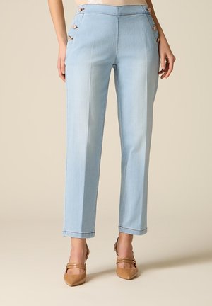 Femme portant un pantalon droit bleu clair avec des boutons latéraux et des escarpins pointus couleur tan sur un fond beige.