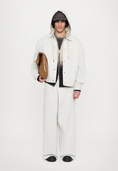 Veste en denim blanche, haut noir, pantalon large blanc avec texture, tenant un sac marron, portant un sweat à capuche et une casquette.