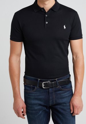 Homme portant un polo noir avec un logo blanc et un jean bleu foncé maintenu par une ceinture en cuir noir avec une boucle argentée.