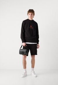 Felpa nera con logo rosso, pantaloni corti neri, sneakers bianche e una piccola borsa nera. Tessuto morbido, vestibilità casual, con polsini e orlo a coste.