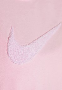 T-shirt rose clair arborant un logo Nike swoosh blanc texturé. Le tissu semble doux avec une texture lisse, mettant en valeur la finition moelleuse du logo.