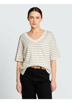 Kobieta o ciemnych włosach, ubrana w biało-beżową koszulkę w paski z dekoltem w serek i krótkimi rękawami, wpuszczoną w czarne spodnie z beżowym paskiem, na neutralnym tle.