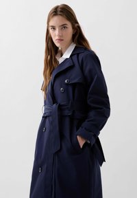 Trench coat bleumarin cu talie prinsa cu cordon, butoane duble si guler clasic. Are un material neted, texturat.