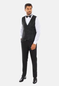 Panciotto a fantasia nera sopra una camicia bianca, abbinato a pantaloni neri e scarpe nere lucide. Presenta un papillon classico.