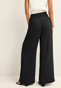 Pantalon noir à jambes larges avec taille haute, doté de poches avant et arrière et d'une texture lisse. Conçu pour une coupe ajustée.