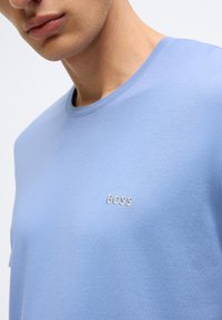 T-shirt de algodão azul-claro com decote redondo, apresentando um discreto logótipo "BOSS" em fio branco bordado no peito. Textura suave.