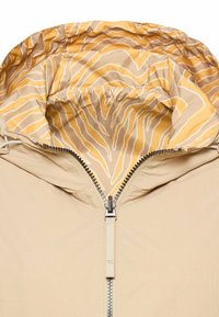 Veste beige avec fermeture éclair à l'avant et capuche doublée d'un tissu à motif rayé zèbre jaune et blanc.