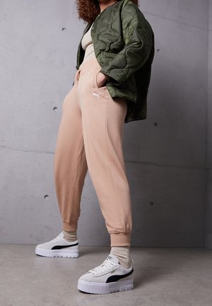 Persoon die lichtroze Puma-sweatpants draagt, witte Puma platform sneakers, beige sokken, een beige top en een groene gewatteerde jas tegen een grijze muur.