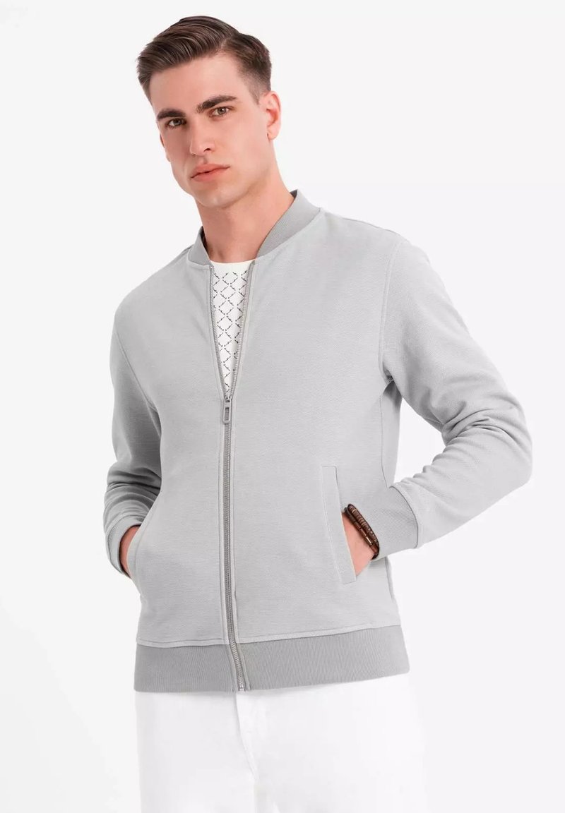 Chaqueta bomber cremosa con cremallera, puños y dobladillo acanalados, que cuenta con dos bolsillos delanteros y una textura suave. El modelo lleva una camiseta estampada debajo.