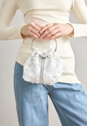 Person holding small white sequin handbag with silver ring handle, wearing cream top and light blue jeans.