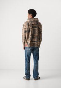 Beige hooded sweatshirt with graphic print, featuring long sleeves and a relaxed fit. Paired with blue jeans and brown shoes.