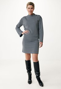 Robe à manches longues en motif géométrique bleu marine et blanc, longueur genou avec un col haut, associée à des bottes noires montantes.
