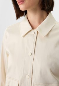 Jasnokremowa kurtka z kołnierzykiem, z przodem zapinanym na guziki i dwiema dużymi przednimi kieszeniami. Gładka faktura materiału.