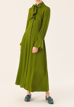 Nainen, jolla on yllään pitkähihainen, oliivinvihreä mekko, jossa on rusetin solmimiskaulus, ja tummanvihreät loaferit, seisoo yksiväristä taustaa vasten.