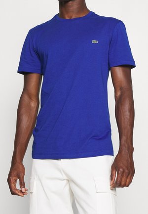 Mężczyzna ubrany w gładką, krótką niebieską koszulkę z małym zielonym logo krokodyla na piersi, zestawioną z białymi spodniami cargo.