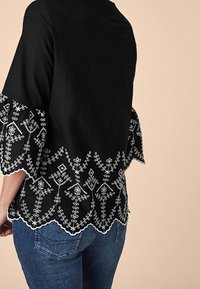 Haut noir à manches trois-quarts avec broderie florale blanche le long de l'ourlet festonné. Porté avec un jean en denim bleu.