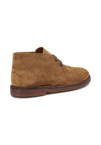 Botas de ante marrón hasta el tobillo con punta redondeada, dos ojales y un tacón bajo, vistas de lado sobre un fondo blanco.