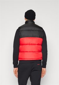 Homme portant une veste matelassée noire et rouge, un bonnet noir et un pantalon noir, debout de dos devant un fond clair uni.