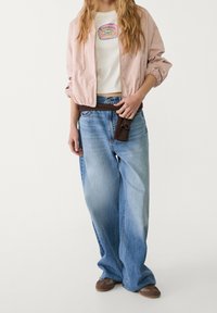 Jasnoróżowa kurtka bomberka, biała koszulka z kolorowym nadrukiem, luźne niebieskie dżinsy i brązowe buty wsuwane. Brązowy pasek związany w talii.