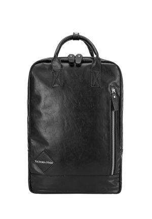 Personne portant une veste en jean bleue et un jean tenant un sac à dos en cuir noir avec des poignées supérieures et une poche avant zippée.