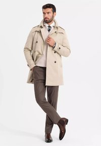 Beżowy dwurzędowy płaszcz trench z pagonami, zestawiony z kremowym swetrem i brązowymi spodniami, dopełniony brązowymi skórzanymi botkami za kostkę.