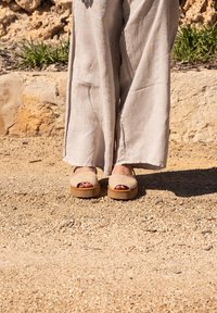 Sandales à plateforme beiges avec bout ouvert, fabriqu�ées en suède doux, dotées d'une large bande supérieure et d'une semelle texturée couleur beige, posées sur un sol sablonneux.