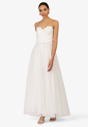 Weißes trägerloses Kleid mit floralem Spitzenoberteil, fließendem Tüllrock und schichtiger Textur. Getragen mit riemchen Sandalen.