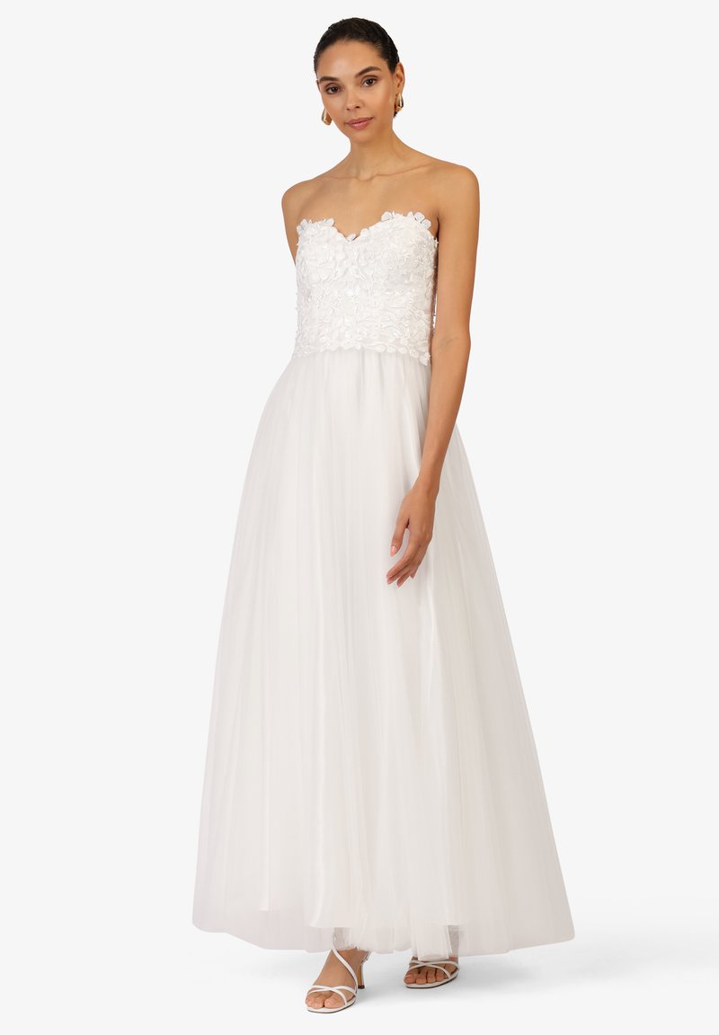 Robe blanche sans bretelles avec un corsage en dentelle florale, jupe en tulle fluide et texture superposée. Portée avec des sandales à lanières.