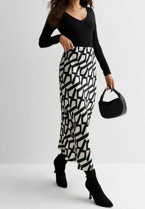 Frau trägt ein schwarzes Langarmoberteil, einen midi Rock mit schwarz-weißem geometrischem Muster, schwarze Stiefel mit Absatz und hält eine kleine schwarze Handtasche.
