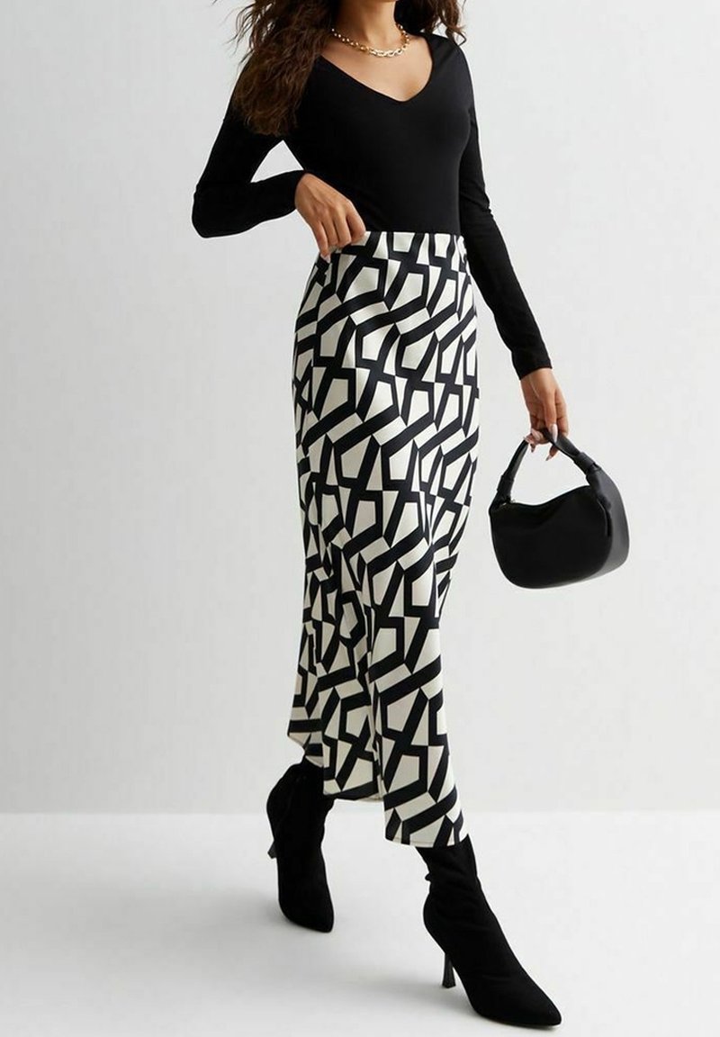 Femme portant un haut noir à manches longues, une jupe midi à motif géométrique noir et blanc, des bottines noires à talons, et tenant un petit sac à main noir.
