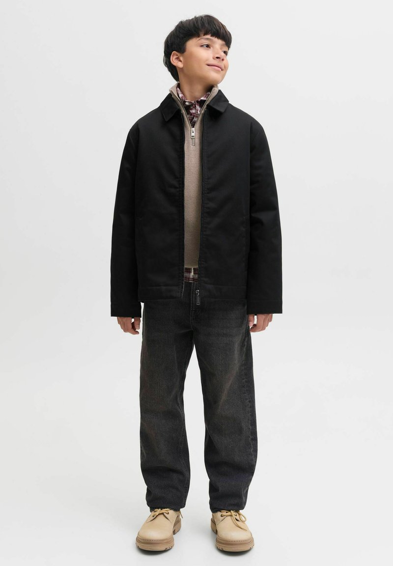 Garçon en veste noire, pull beige à fermeture éclair, chemise à carreaux, jean foncé et chaussures beiges, debout devant un fond clair uni, regardant sur le côté.