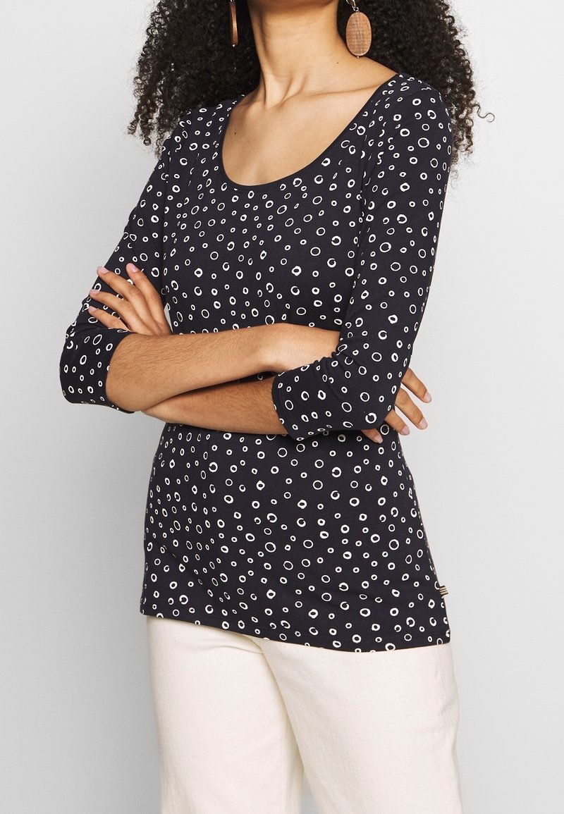 Tmavě modrý top s dlouhými rukávy a bílými kulatými vzory; kulatý výstřih; měkká tkanina; přiléhavý střih; nošený s světle zbarvenými kalhotami.