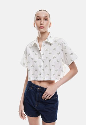 Camicetta bianca cropped con motivo floreale verde, maniche corte, colletto a punta, texture di tessuto a strati; abbinata a pantaloni shorts in denim blu scuro.