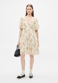 Κρεμ φόρεμα με δαντέλα σε λουλουδάτο σχέδιο, με φουσκωτά μανίκια, μήκος μέχρι το γόνατο και V-λαιμόκοψη· συνδυασμένο με μαύρη τσάντα, λευκές κάλτσες και μαύρα μποτάκια.