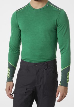 Homme portant une chemise verte à manches longues avec des détails vert foncé et jaune sur les manches et un pantalon cargo noir, debout avec les bras détendus.