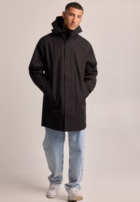 Impermeable ligero negro con cuello alto, cierre de botones y bolsillos laterales, combinado con jeans anchos de color azul claro y zapatillas blancas.